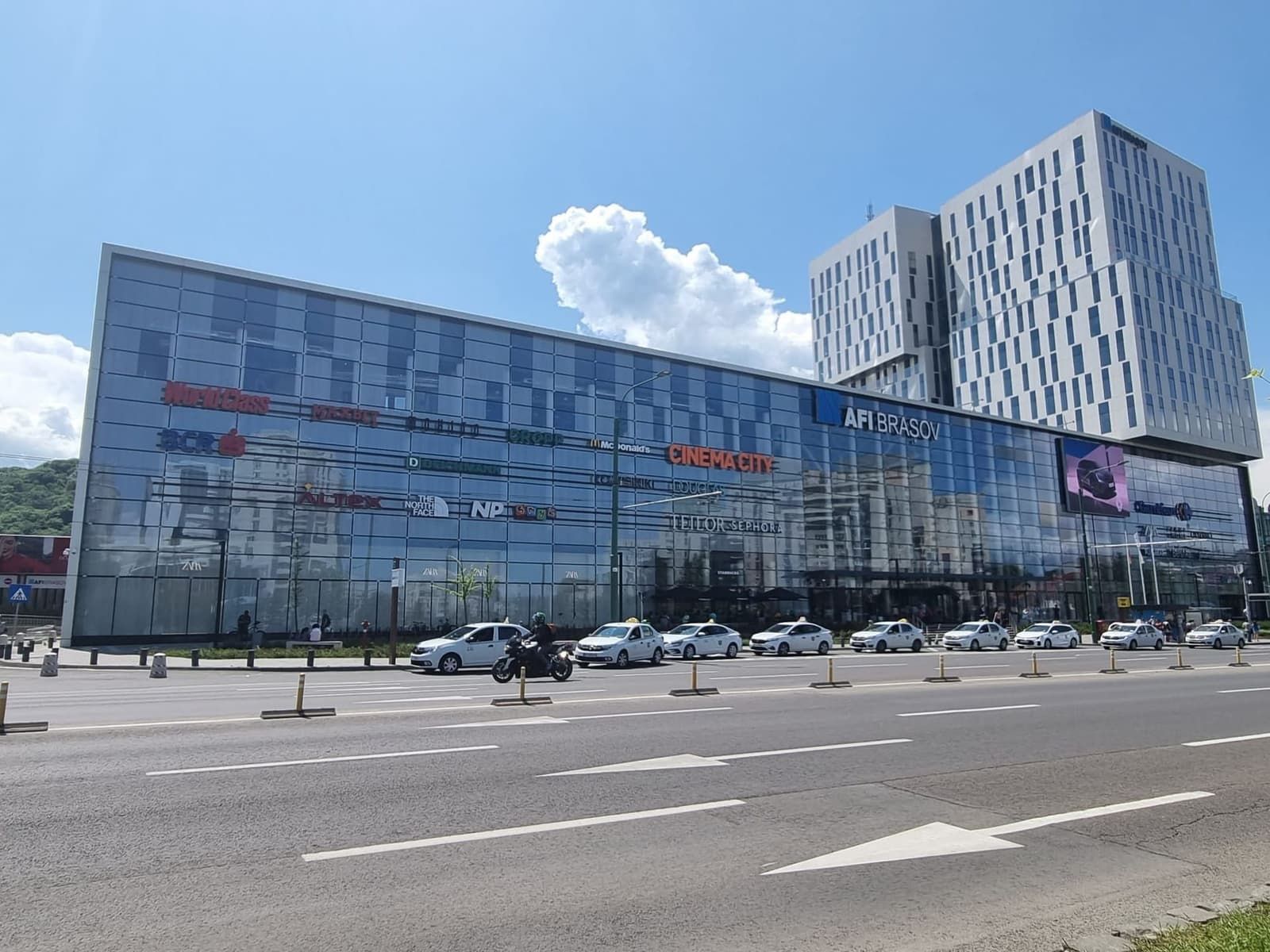 The glass front of AFI Brașov on Bulevardul 15 Noiembrie, with shop signs, a row of taxis and the office tower behind