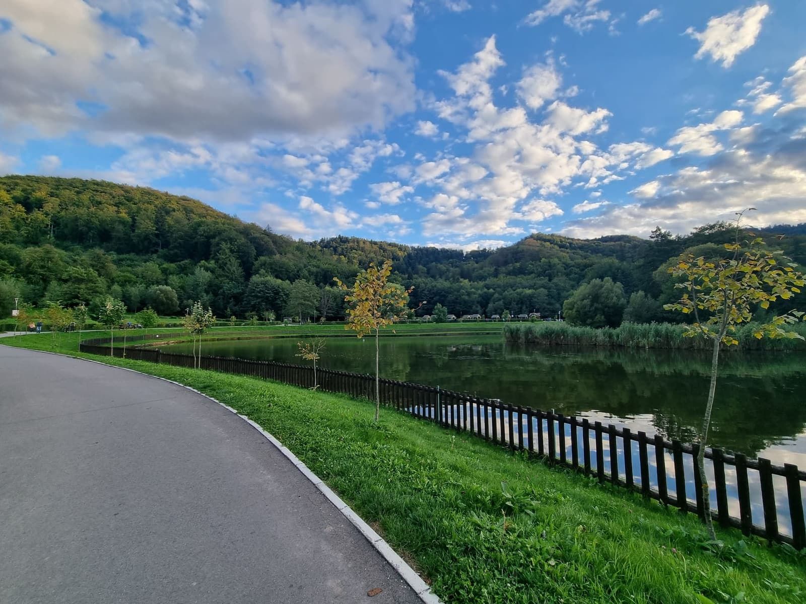 A paved path beside the lake at Noua, with forested hills behind