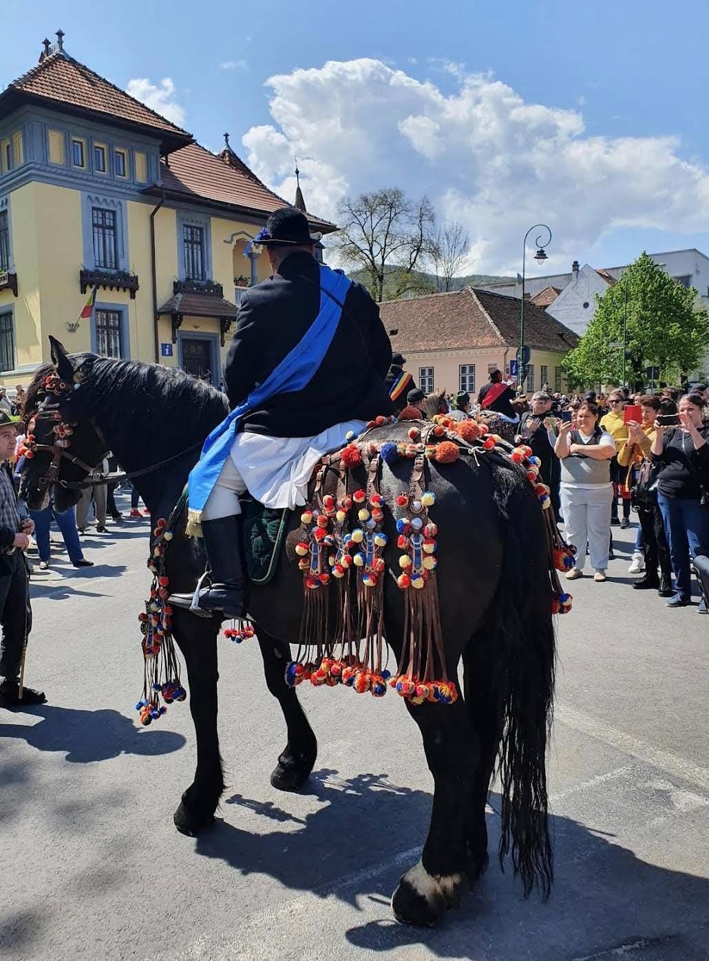 Jun călare pe un cal negru împodobit cu ciucuri roșii, galbeni și albaștri, privit de mulțime